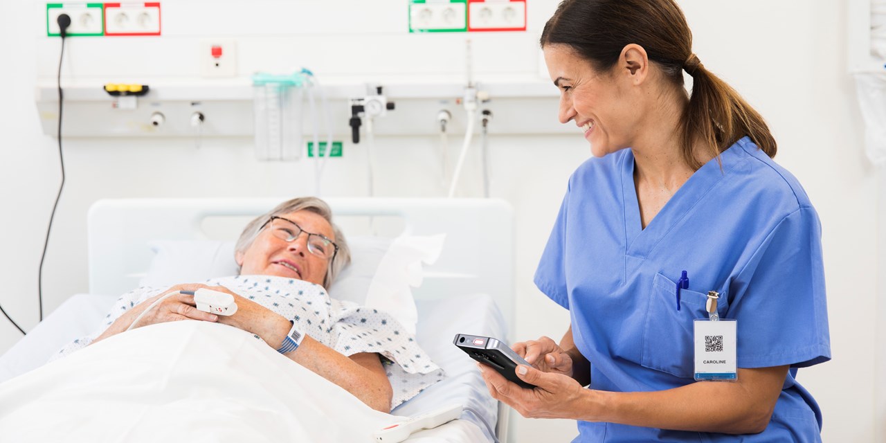 Sitting caregiver holds smart phone device while conversating with a monitored patient in bed