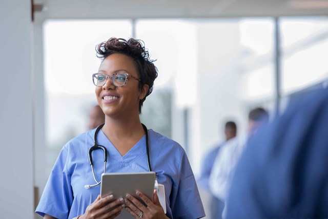 A smiling nurse in scrubs is holding a tablet