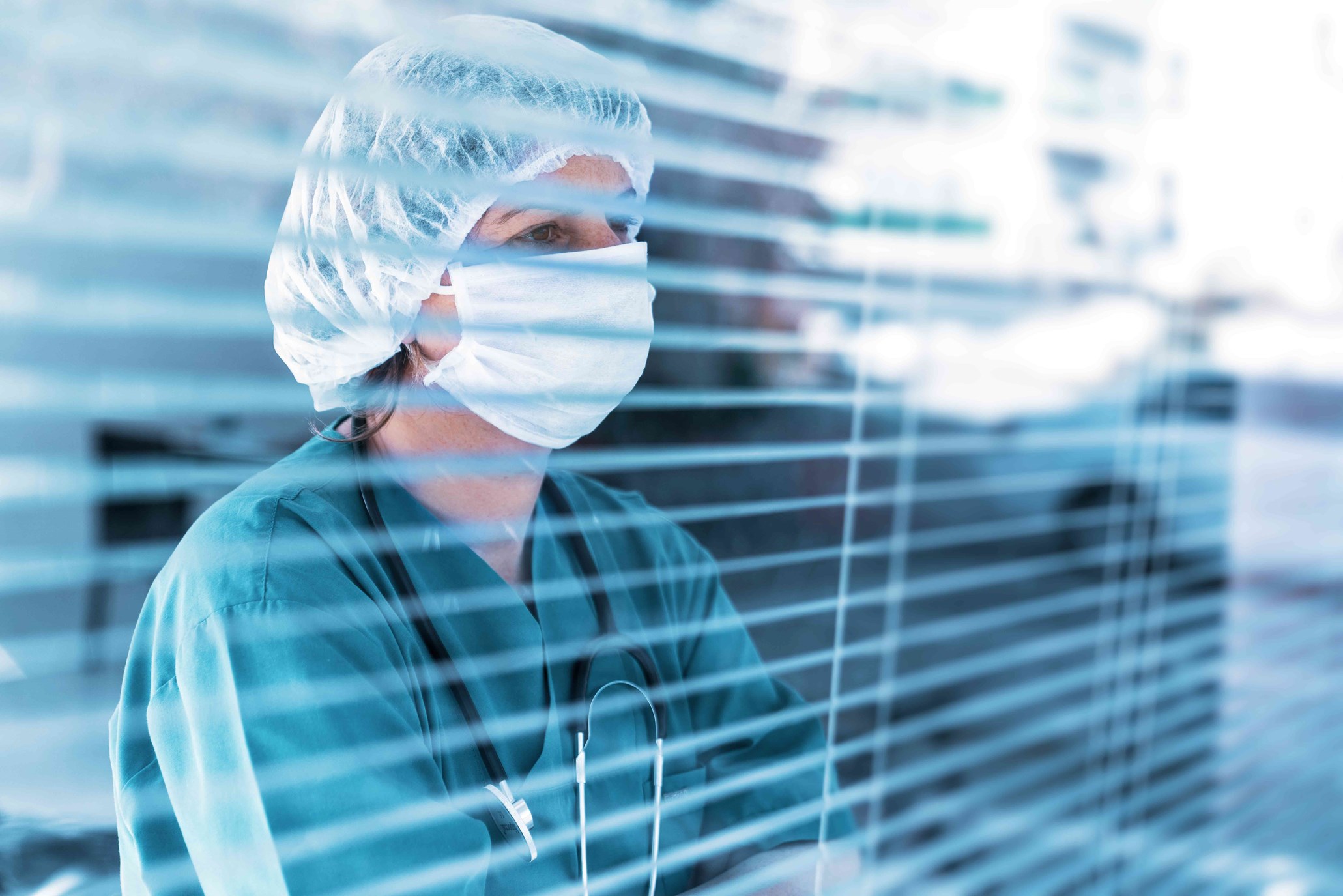 Caregiver wearing scrubs, a stethoscope, a surgical mask, and a surgical cap looking through a window