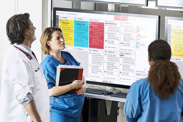 Two female nurses in blue scrubs and a male doctor in a white coat in front of a dashboard displayed on a big screen
