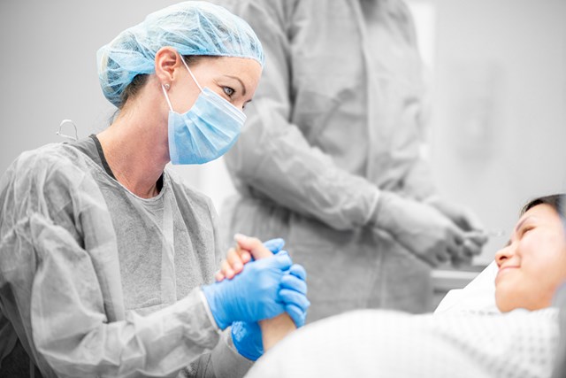 Female nurse in PPE, holding the hand of a pregnant woman in a delivery room