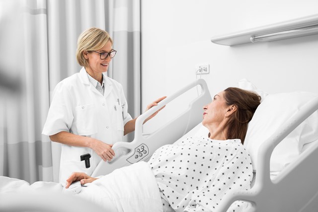 Female medical professional by the bedside of female patient, smart phone device can be seen in the pocket of the caregiver's white coat