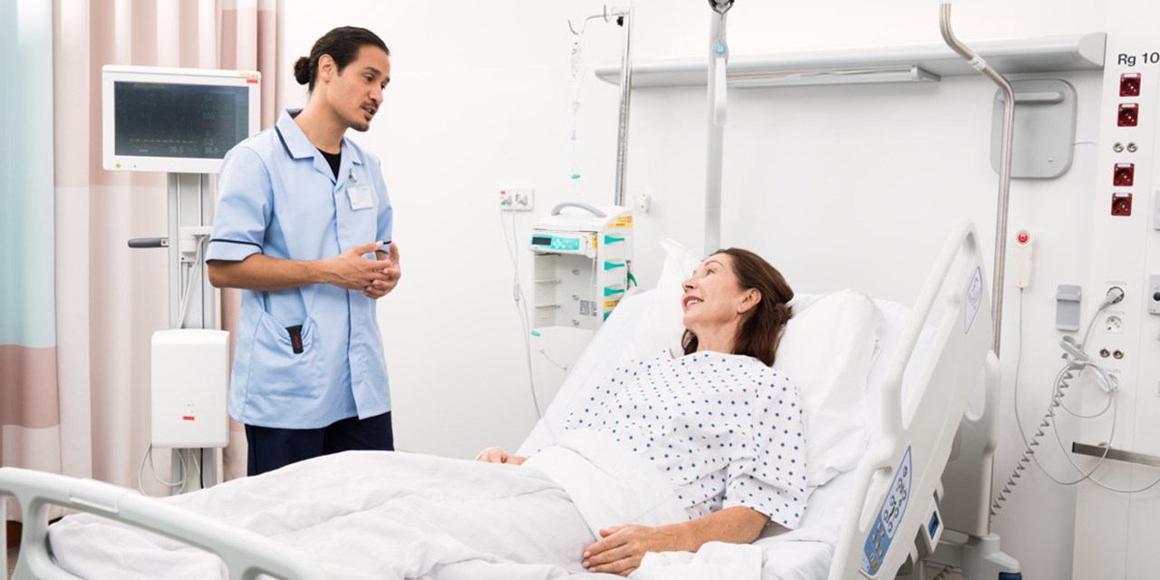 A standing male nurse interacts with a female patient in bed