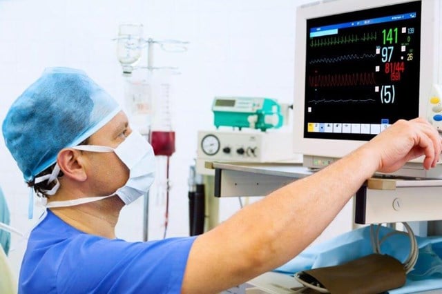 Clinician wearing scrubs, a surgical cap and surgical mask looking at monitor