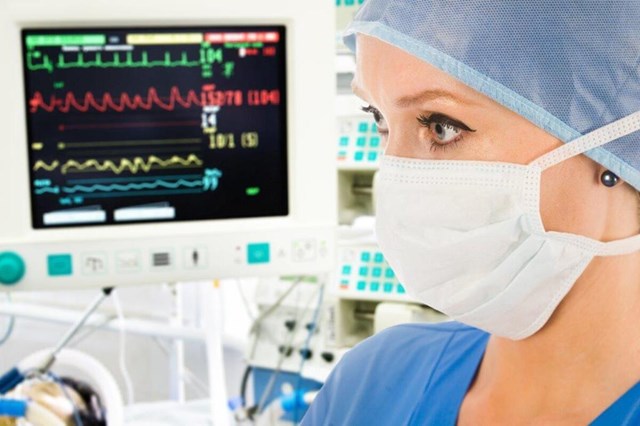 OR nurse wearing scrubs, a surgical mask, and a surgical cap with blurred monitor in background