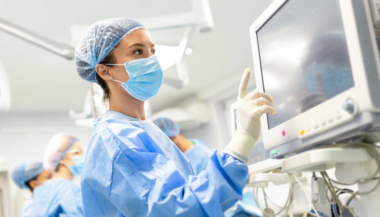 Close-up of female medical personnel in PPE looking at a monitor, there are other members of the staff in the blurred background