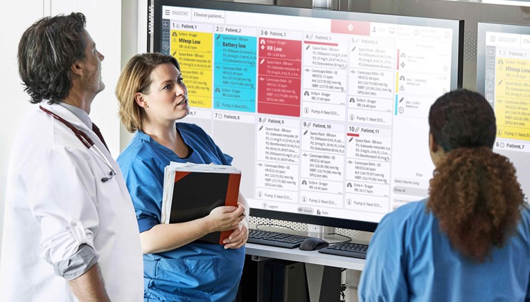 Two female nurses in blue scrubs and a male doctor in a white coat in front of a Digistat dashboard displayed on a big screen