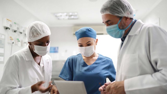 Three doctors in scrubs, surgical masks and a surgical cap discussing something they are seeing on a tablet