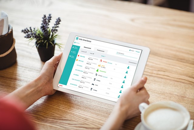 Close-up of hands holding a tablet displaying a resident overview screen from Ascom SmartSense, over a desk.