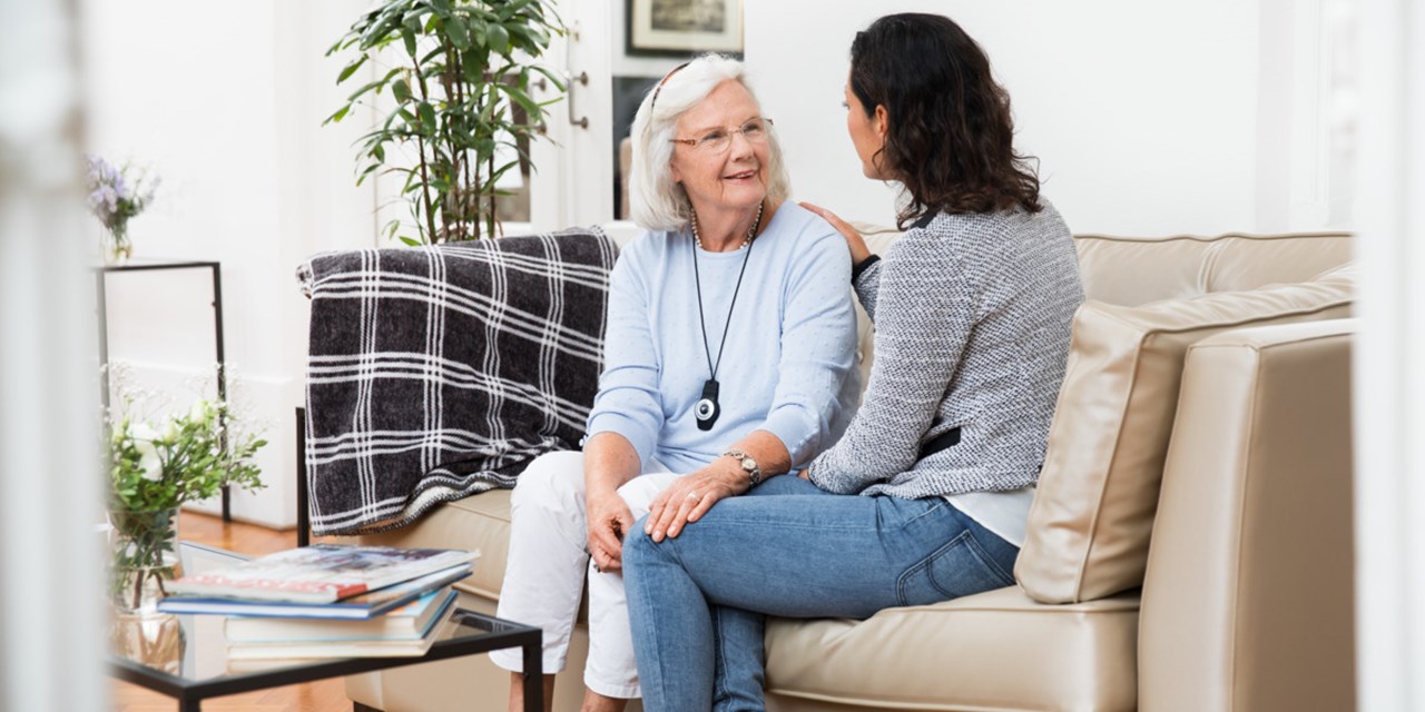 An elderly woman sits on a sofa and interacts with a female visitor; the resident wears a teleCARE pendant.